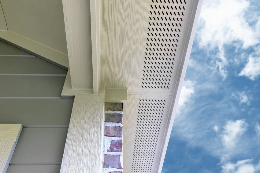 Close-up view of a white vented soffit under a roof edge with a partly visible brick wall and blue sky with clouds in the background.