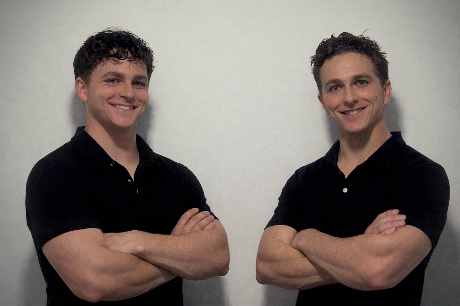 Two young men standing side by side with arms crossed, both wearing black shirts and smiling against a plain light background.