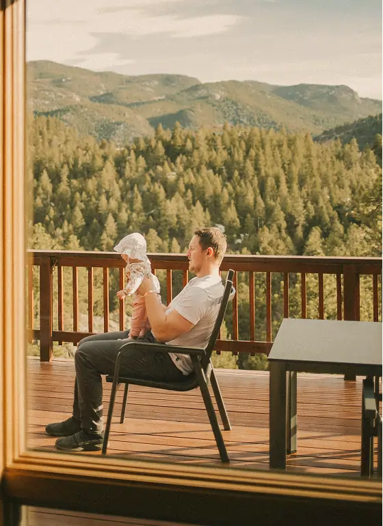 Man sitting on a chair holding a baby on a wooden deck with a forested mountain background.