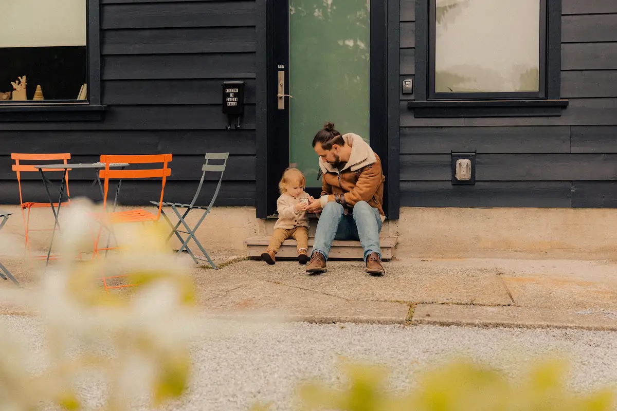 Man and young child sitting on steps outside black house with orange and gray chairs nearby.