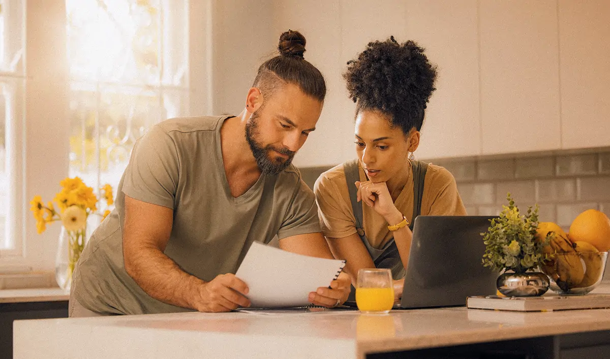 Man and woman reviewing documents together at a kitchen counter with a laptop and a glass of orange juice nearby.