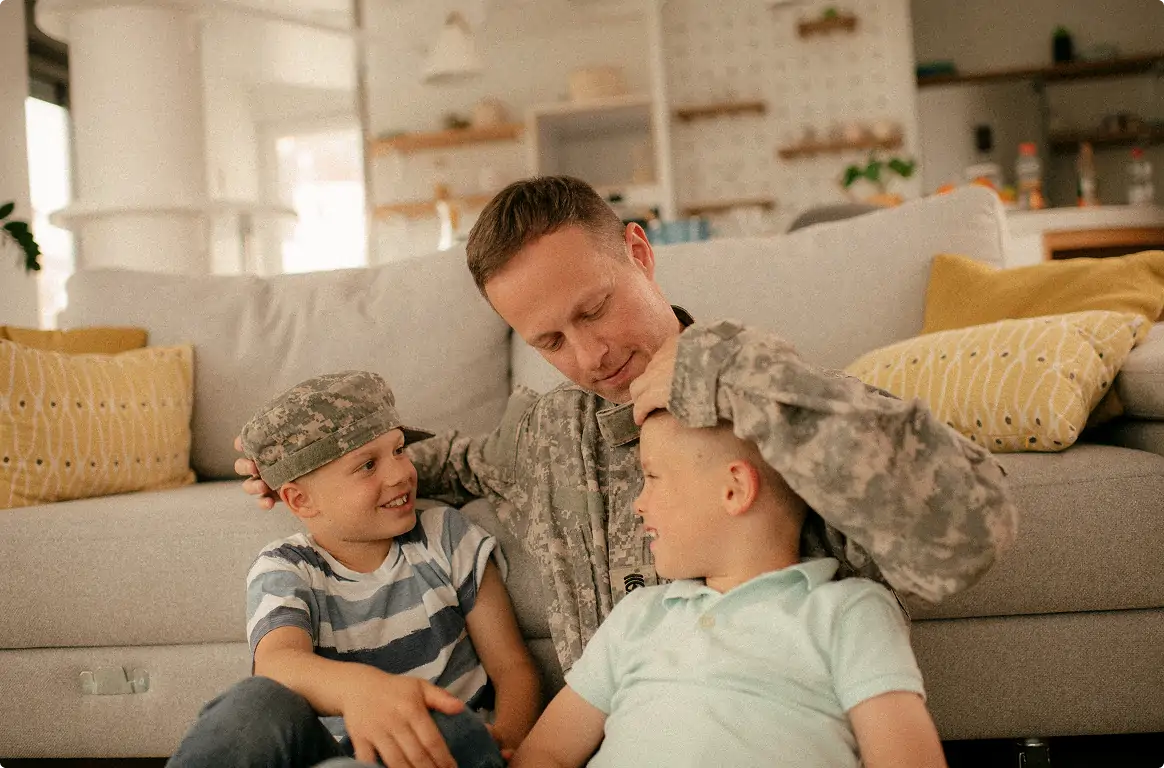 Man in military uniform sitting on the floor with two smiling young boys, one wearing a military cap, in a cozy living room.