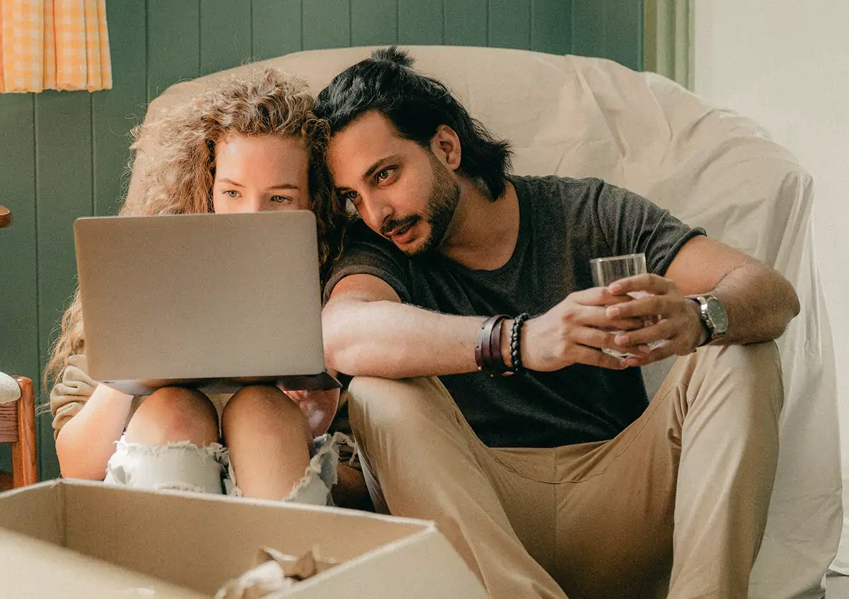 A couple sitting on the floor looking at a laptop, with a cardboard box in front of them.