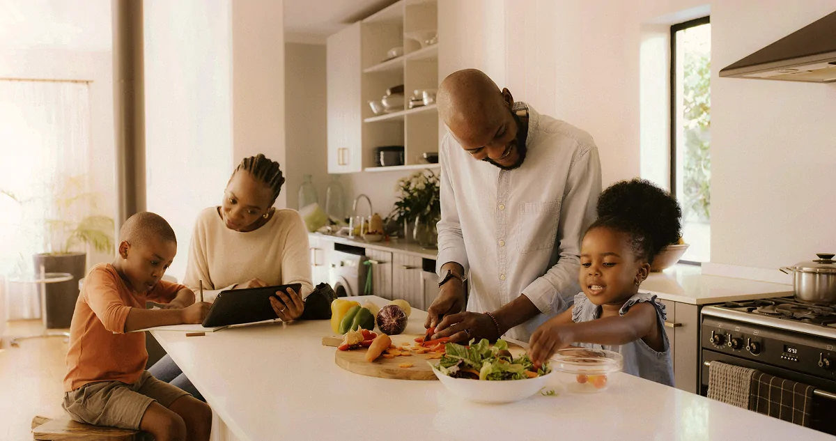 Family in kitchen with father cutting vegetables, mother using a tablet, son writing, and daughter reaching for salad.