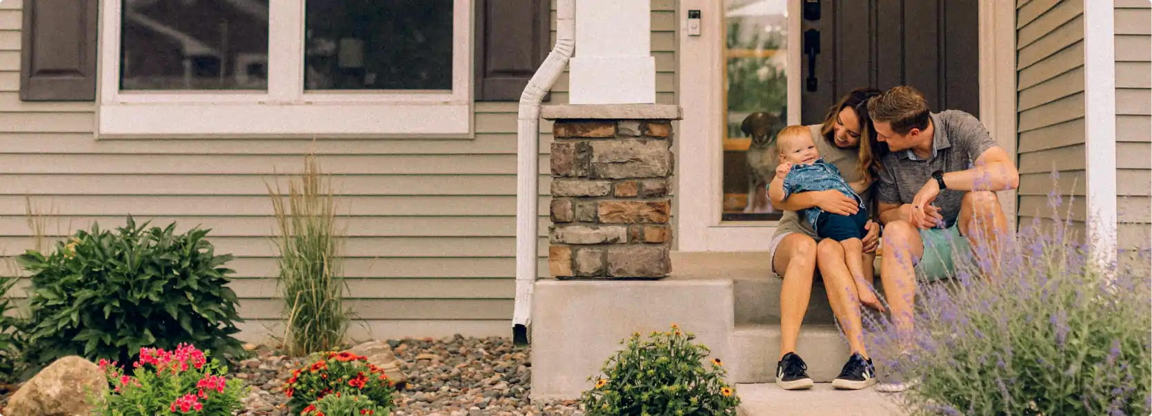 Parents sitting on front porch steps holding their baby with a dog visible through the glass door.