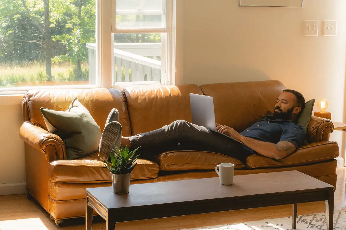 Man reclining on a brown leather couch using a laptop in a sunlit room with a coffee mug and potted plant on a wooden table.