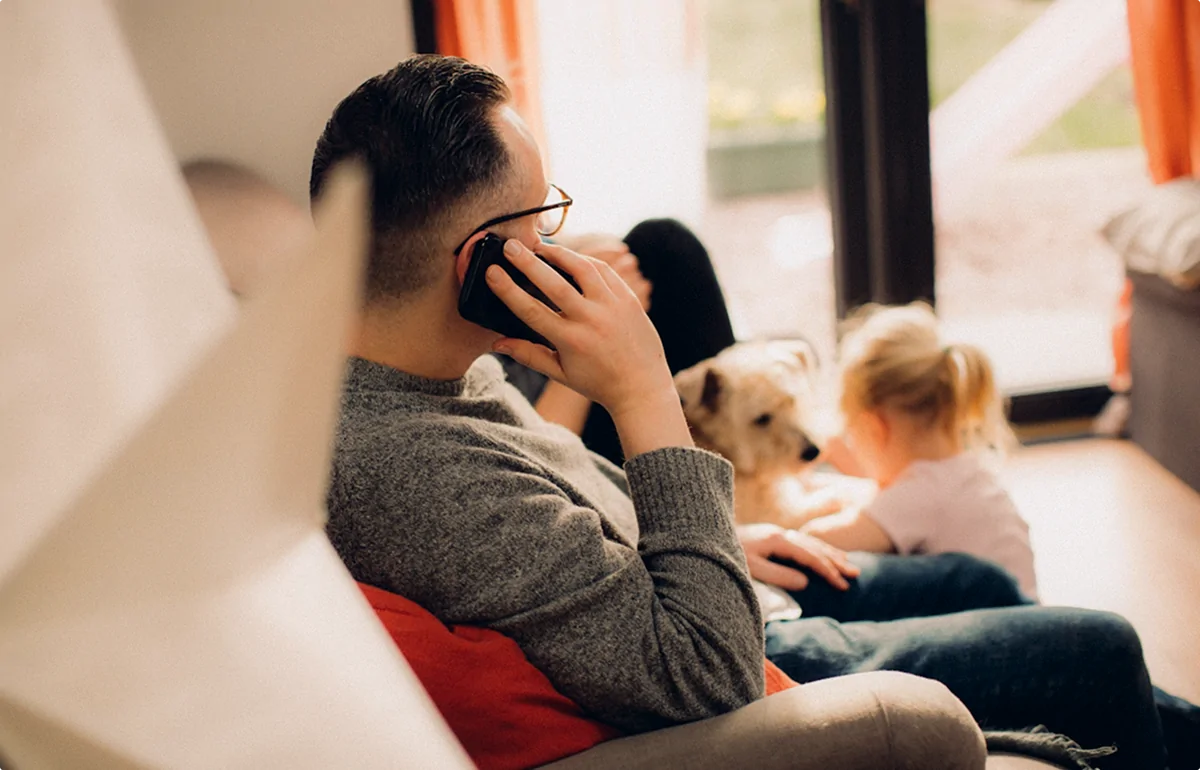 Man sitting on a couch talking on a phone while a young girl plays with a dog in the background.