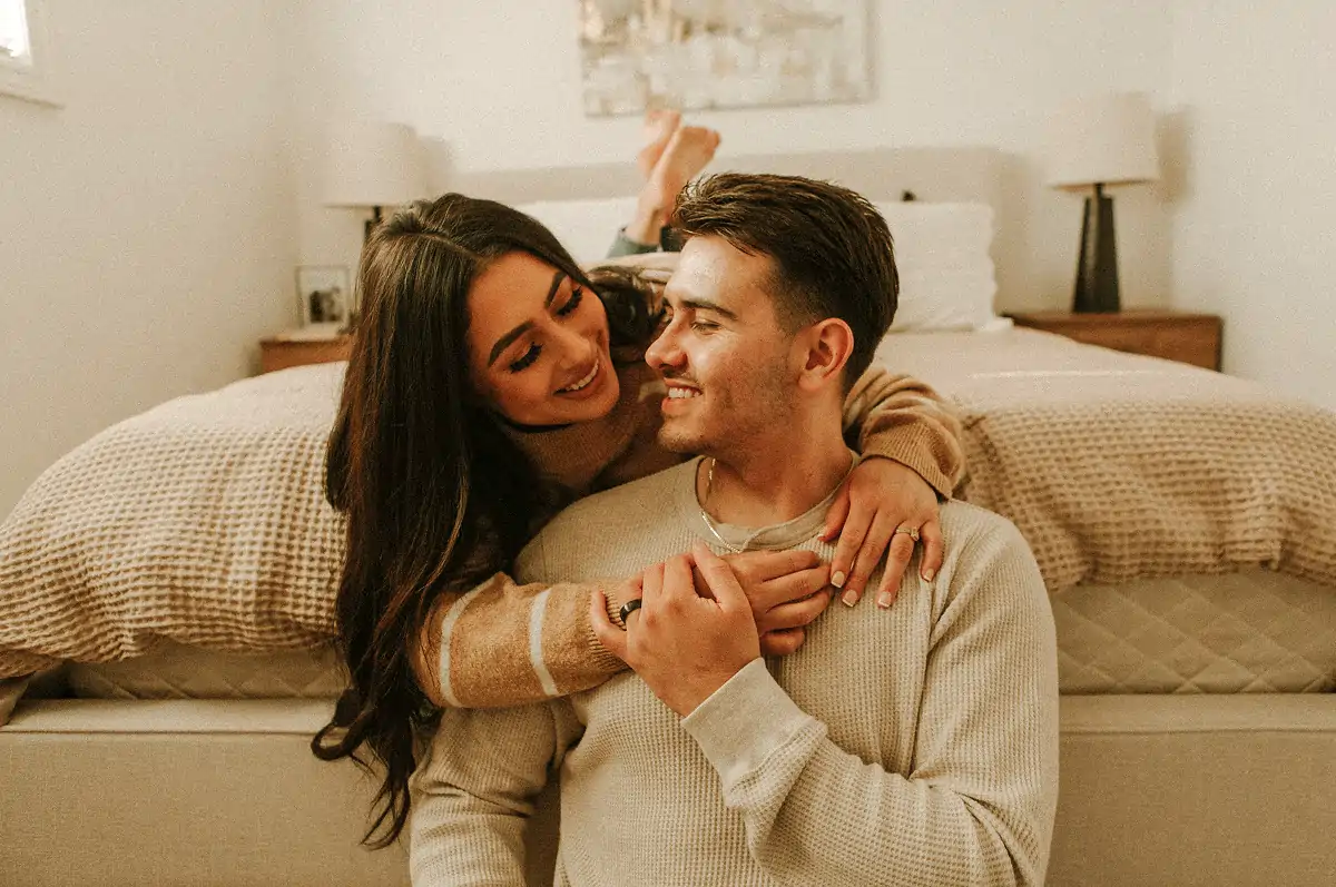 Smiling couple embracing affectionately in a cozy bedroom with beige decor.