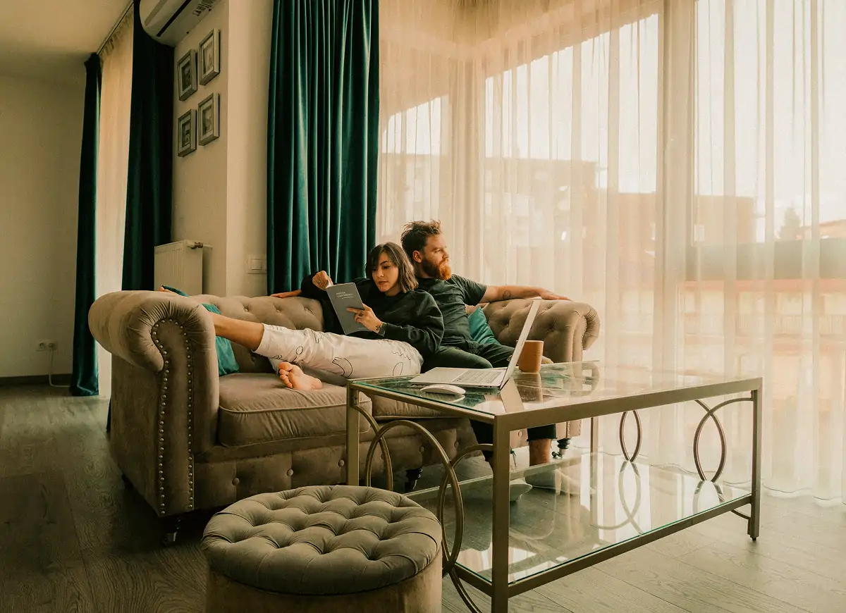 Couple relaxing on a beige sofa in a sunlit living room, the woman reading a book and the man sitting behind her with a laptop on a glass coffee table.