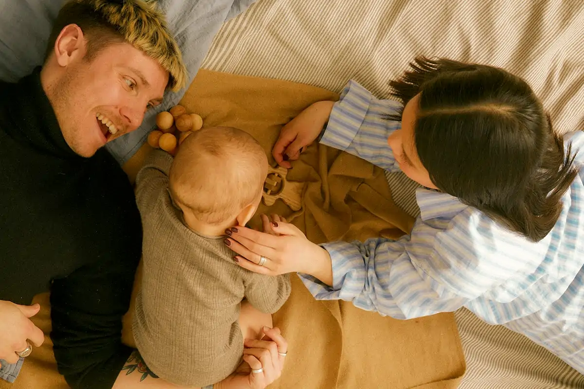 Young couple lying on a bed playing with their baby who is reaching for wooden toys.