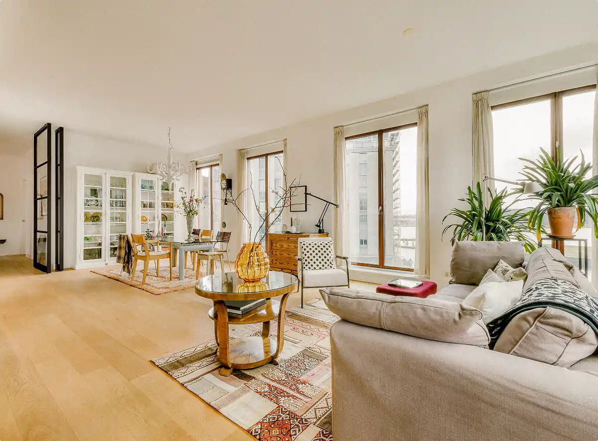 Bright living room with beige sofa, large windows, green plants, round wooden coffee table, and dining area with glass cabinet.
