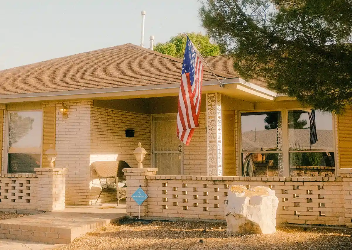 Single-story brick house with an American flag on the porch and a large rock in the front yard.