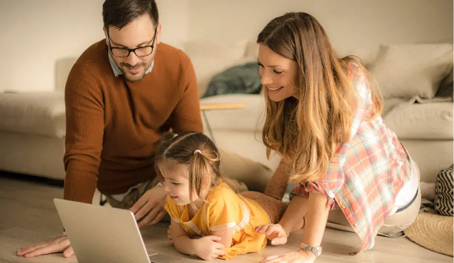Smiling parents with their young daughter lying on floor watching a laptop together in a cozy living room.