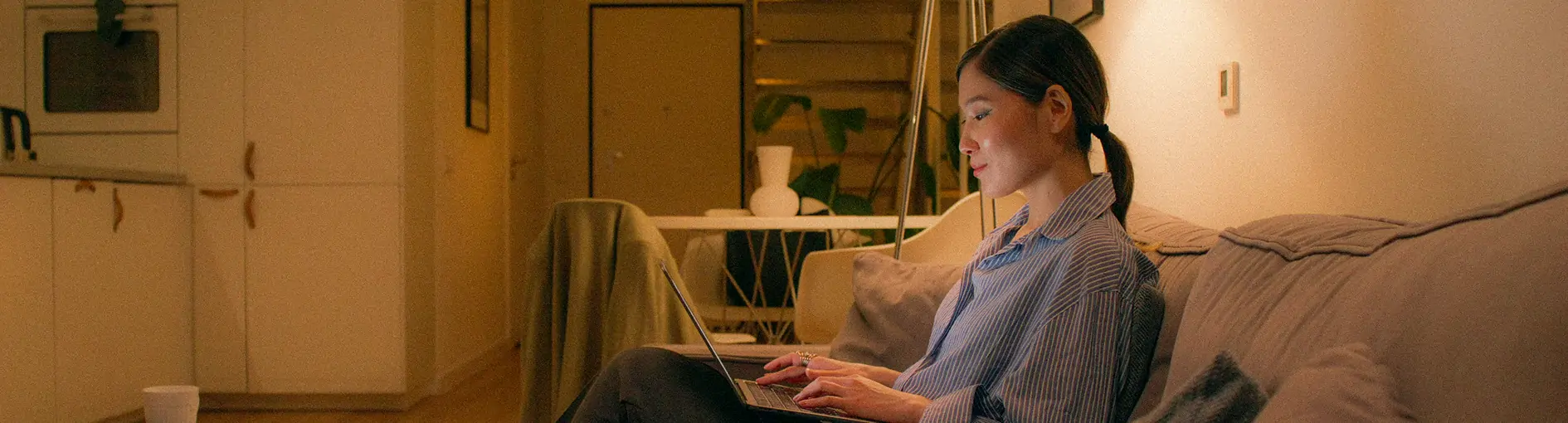 Woman sitting on a couch in a cozy living room, working on a laptop in soft warm lighting.
