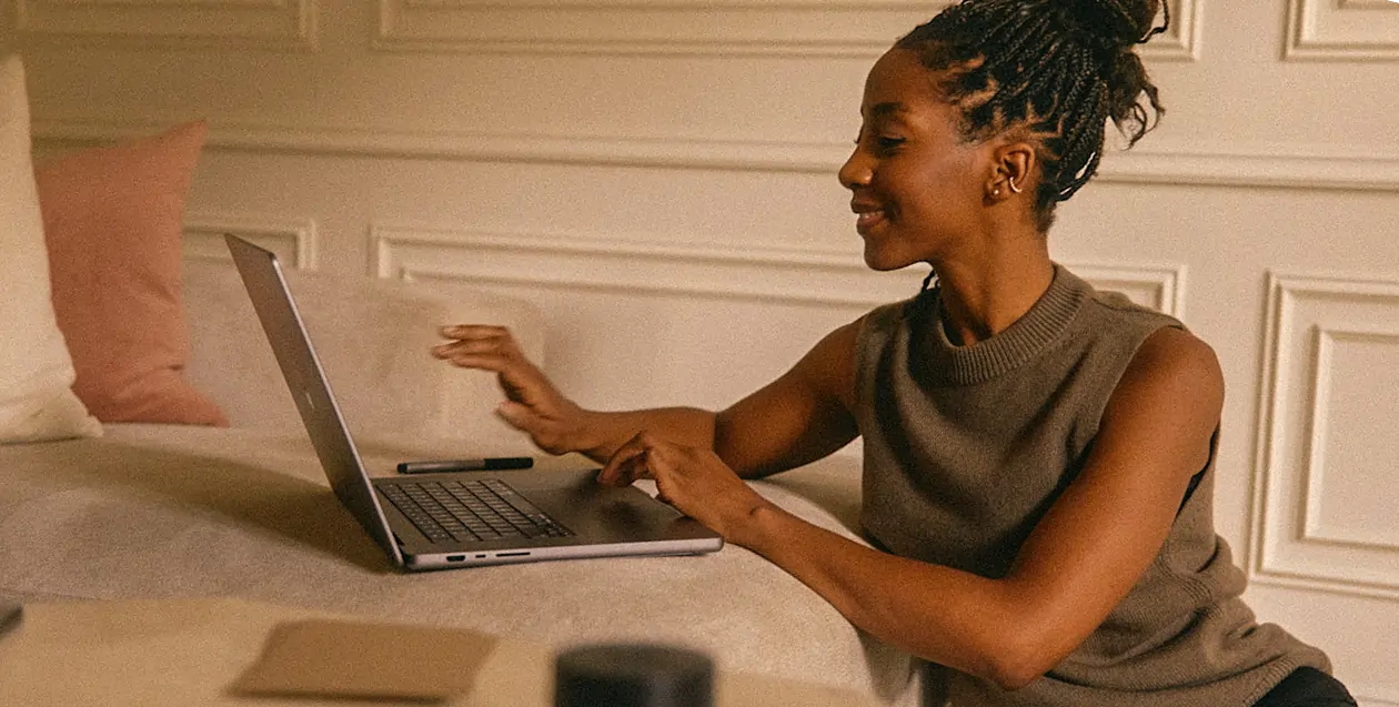 Smiling woman with braided hair using a laptop on a beige couch with cushions in a softly lit room.