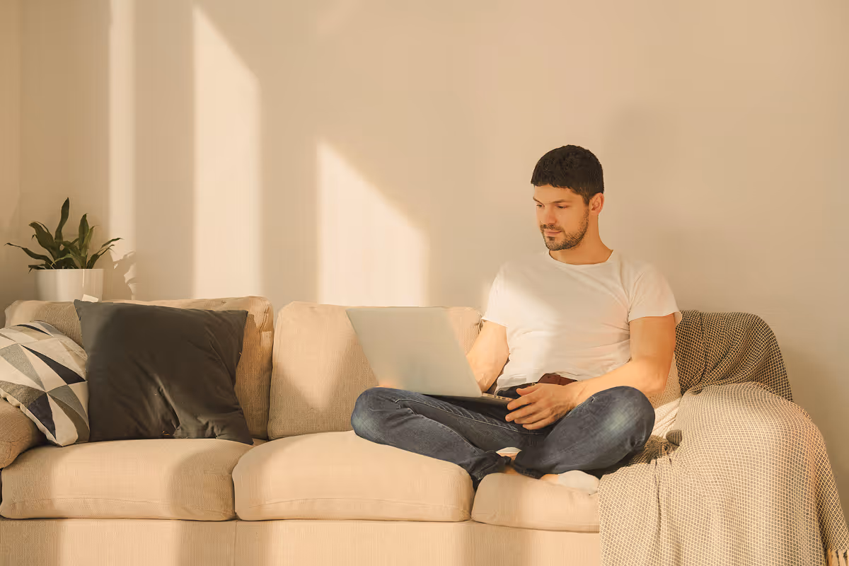 Man in white t-shirt and jeans sitting cross-legged on a beige couch working on a laptop with sunlight streaming on the wall behind.