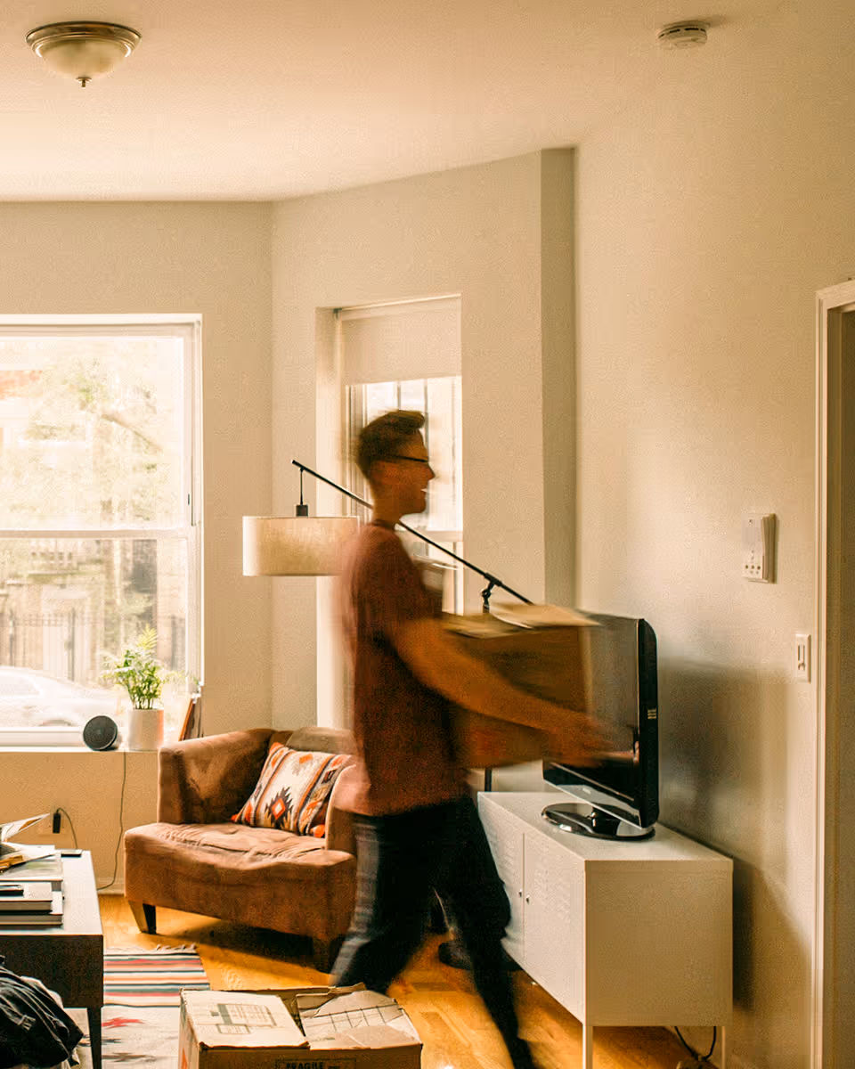 Blurred image of a man carrying a cardboard box in a bright living room with a couch, lamp, and TV.