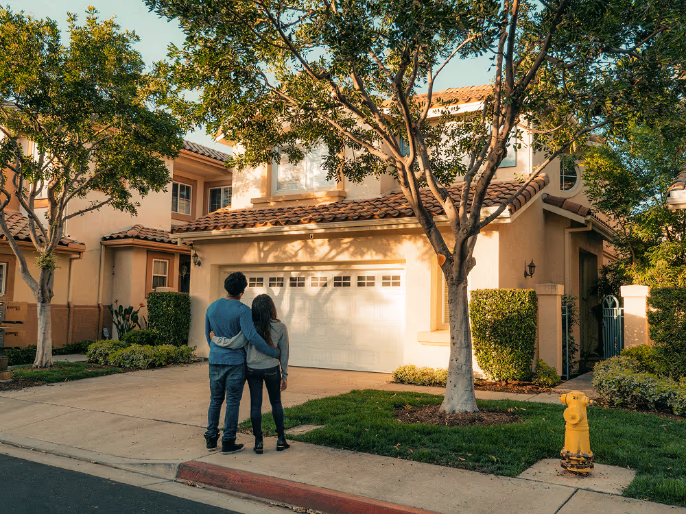 Couple embracing while standing on a sidewalk and looking at a two-story suburban house with a white garage door.