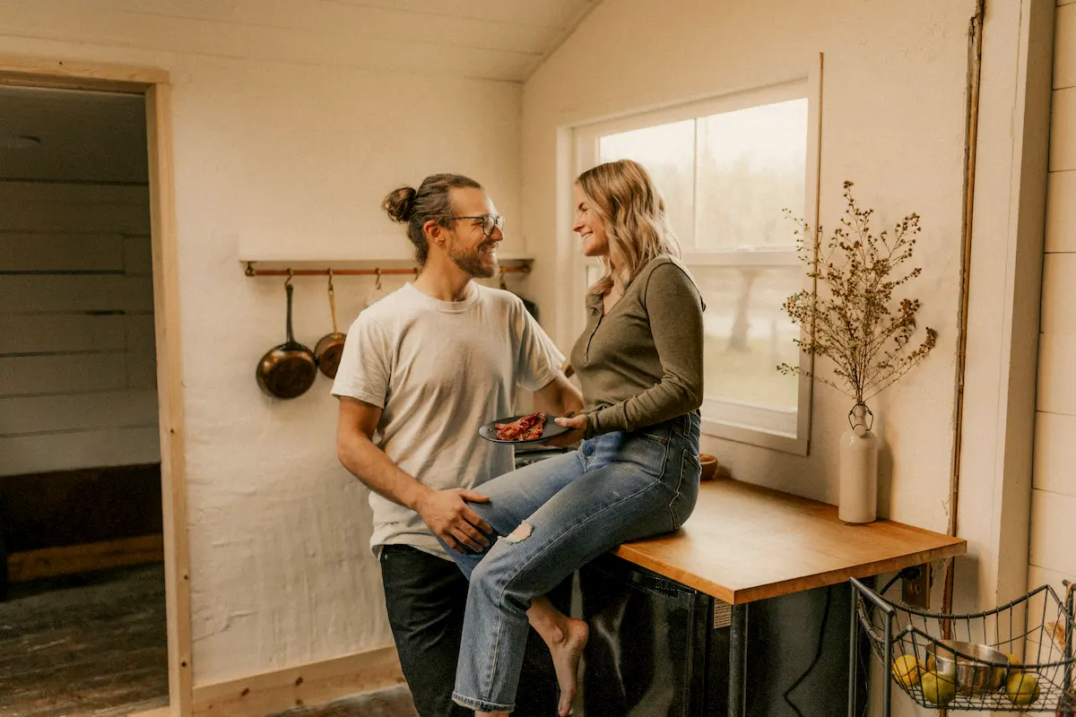 A man and woman smiling at each other in a cozy kitchen, with the woman sitting on a wooden countertop holding a plate with food.