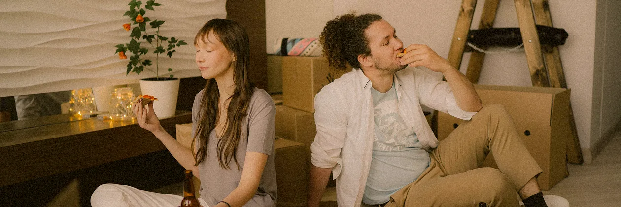 A couple sitting on the floor in a room with moving boxes, enjoying slices of pizza with relaxed expressions.