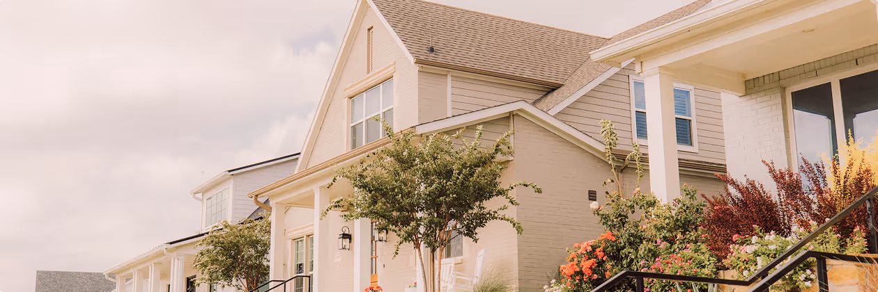 Row of beige houses with large windows, front porches, trees, and colorful flowering bushes under a cloudy sky.