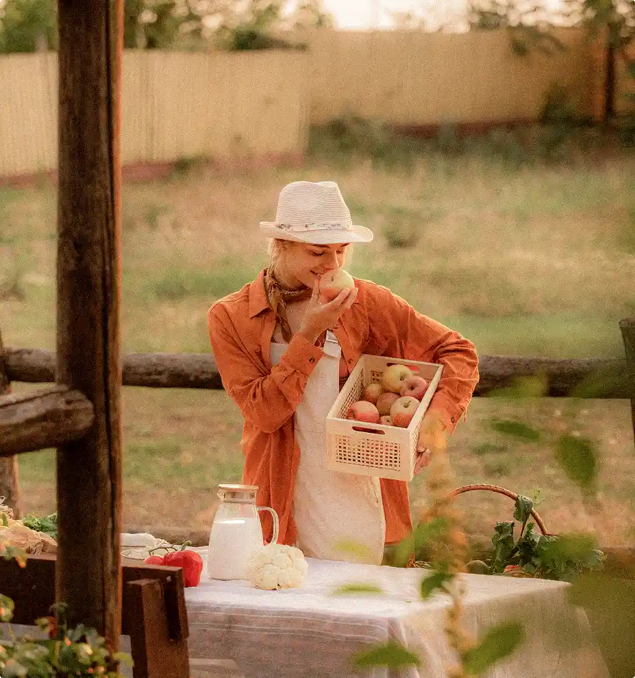 Woman in a hat and orange shirt holding a basket of apples and smelling one by a table set outdoors with vegetables and milk.