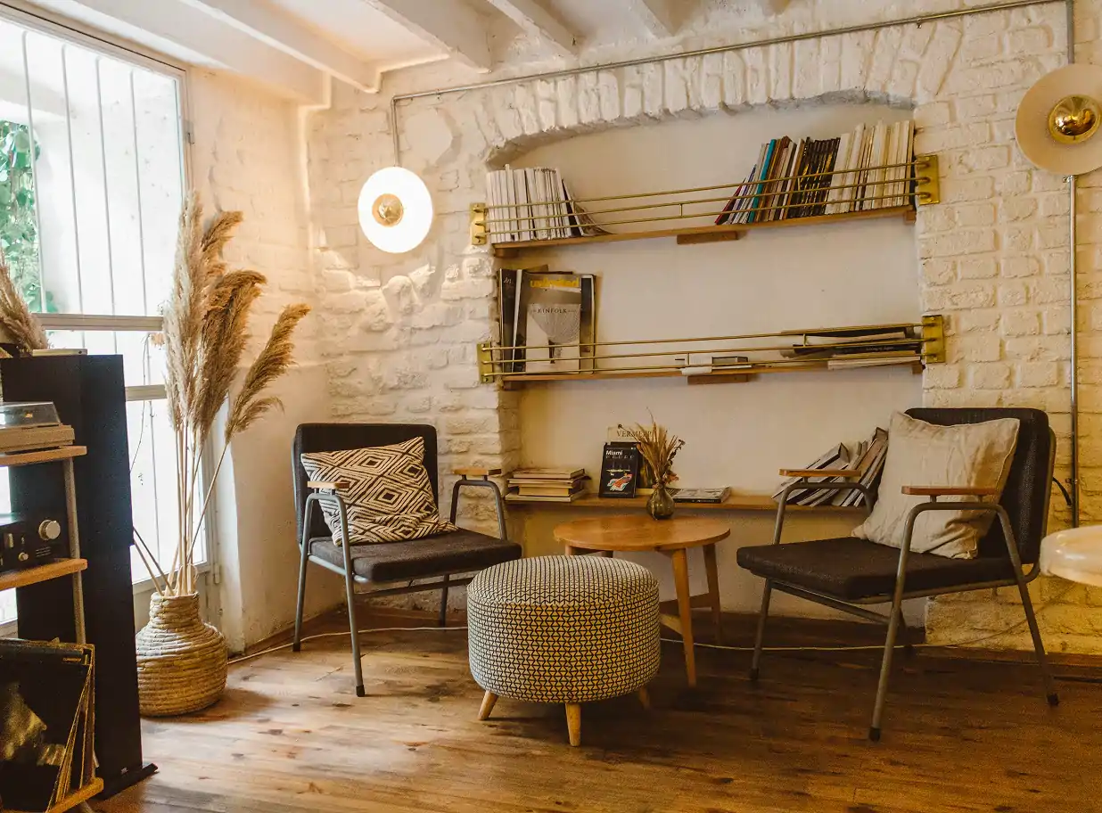 Cozy retro-style sitting area with two black armchairs, a patterned round ottoman, wooden side table with a small vase, and shelves with books on a white brick wall.