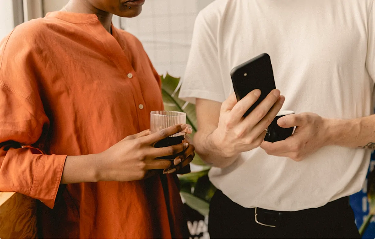 Two people standing and holding drinks, one showing something on a smartphone to the other.