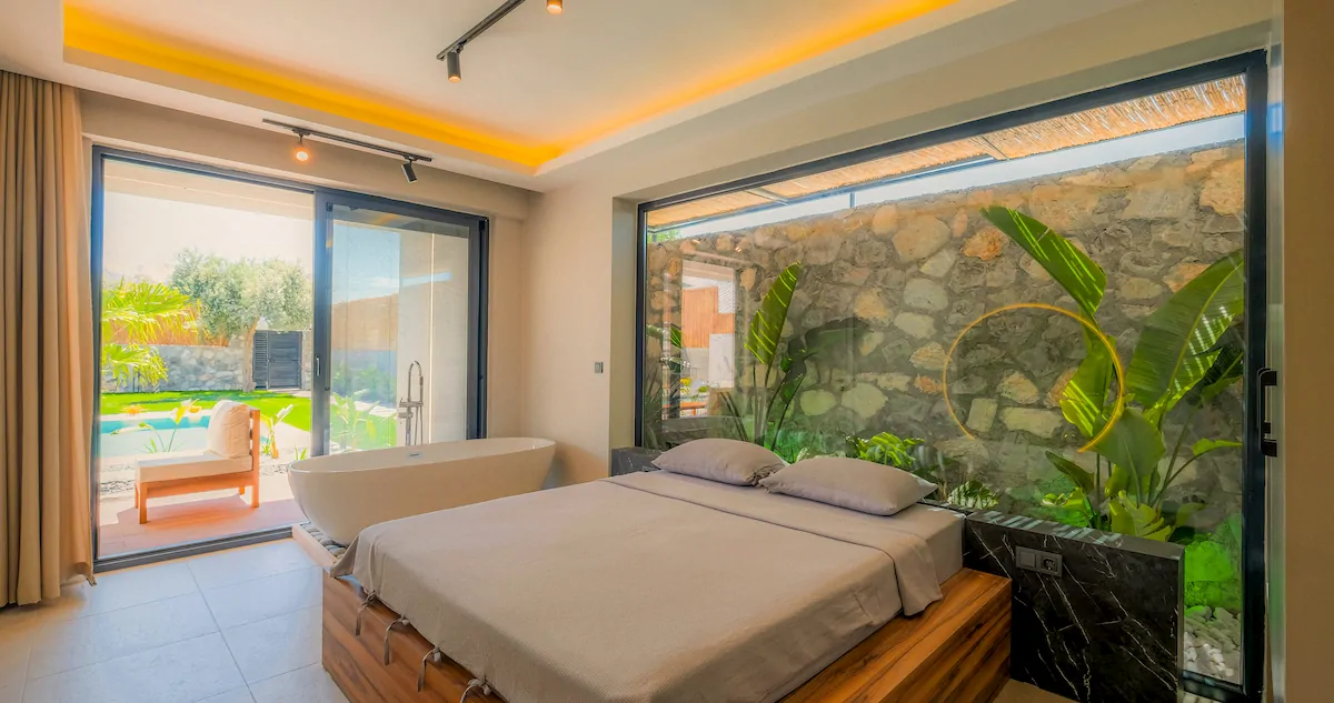 Modern bedroom with wooden platform bed, large pillows, indoor plants behind glass wall, and a freestanding bathtub near sliding doors opening to a garden with a pool.