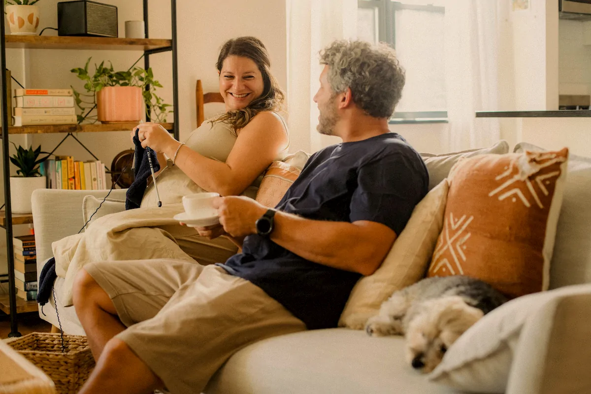 Smiling couple seated on a beige couch; woman is knitting while man holds a cup, with a small dog resting on the couch.