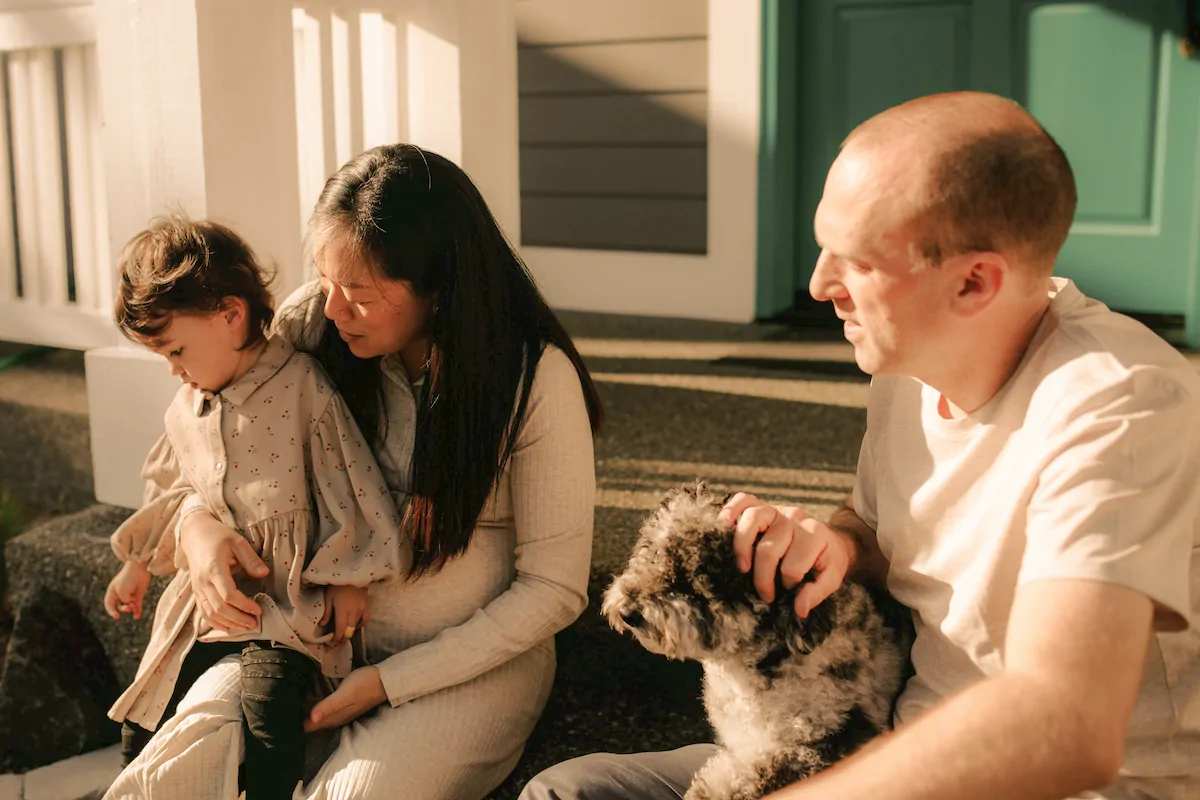 A couple with a young child sitting on steps outside, the woman holding the child and the man petting a small dog.
