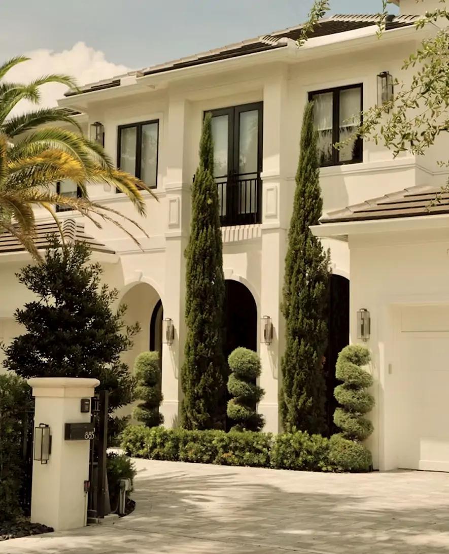Elegant white two-story villa with tall, manicured trees and a paved driveway.