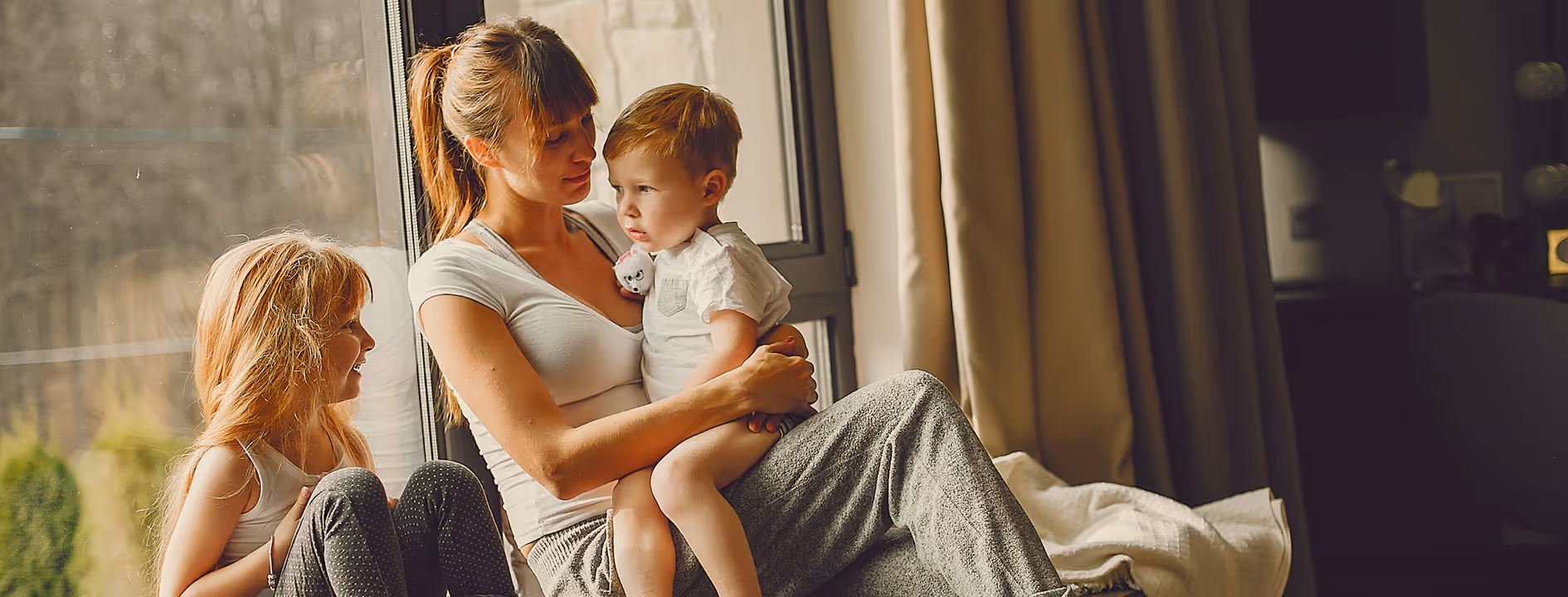 Mother sitting by a window holding a young boy, while a young girl sits nearby looking at them.