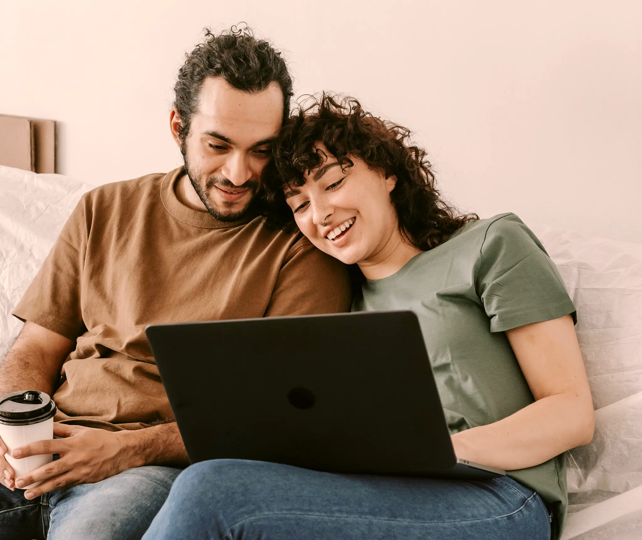 A couple with curly hair sitting close together on a bed, smiling while looking at a laptop.