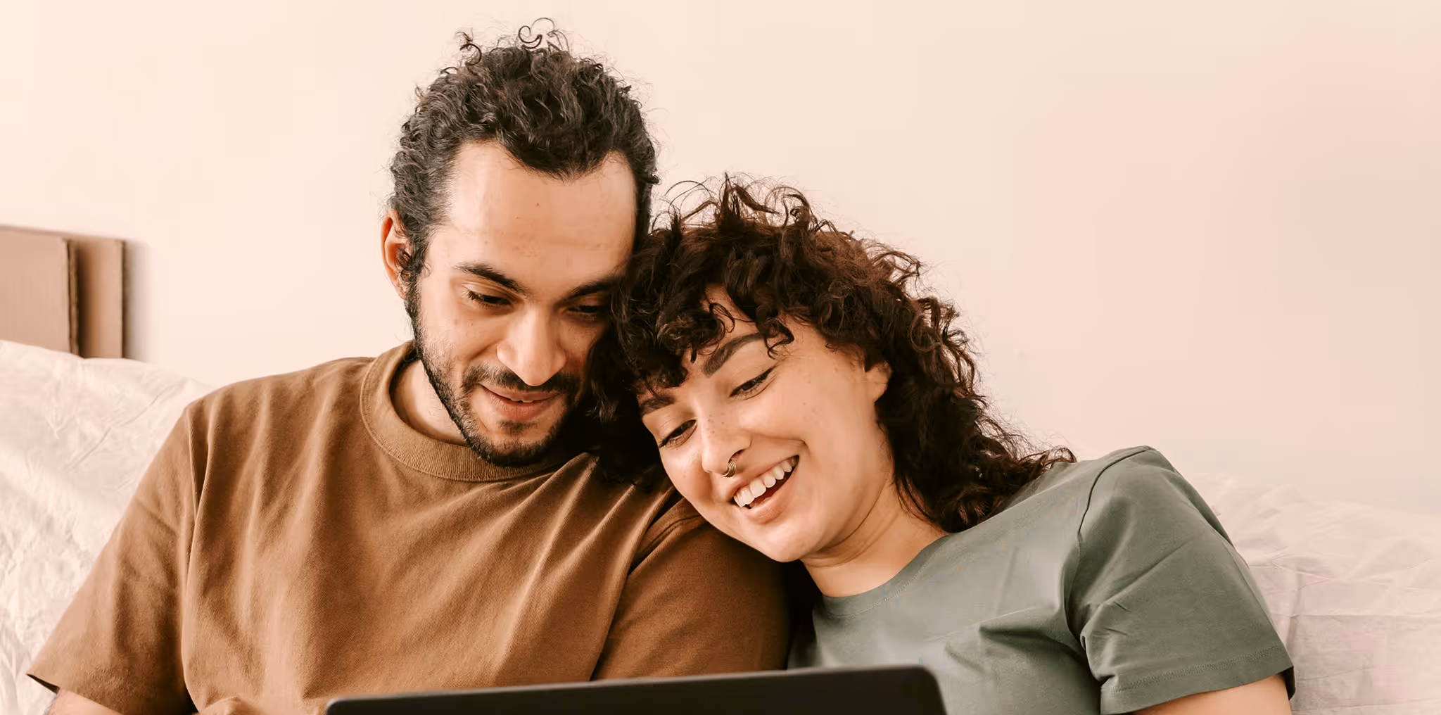 A couple with curly hair sitting close together on a bed, smiling while looking at a laptop.