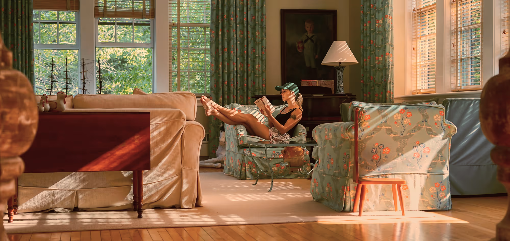 Woman wearing a cap reads a book while reclining on a floral armchair in a sunlit living room with large windows and patterned curtains.
