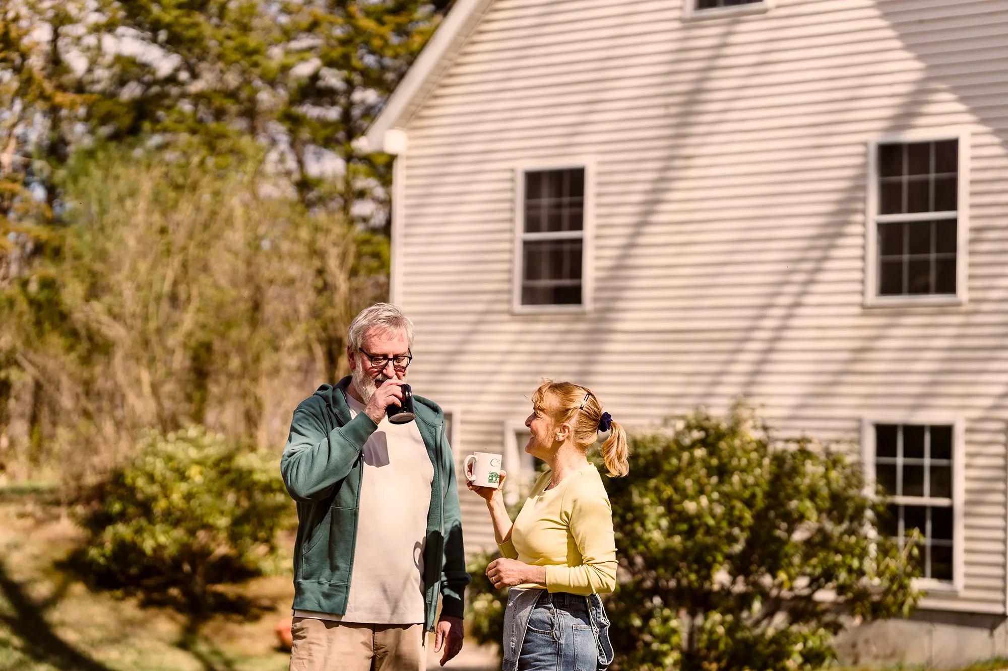 Older couple enjoying drinks and smiling outdoors near a white house with trees in the background.
