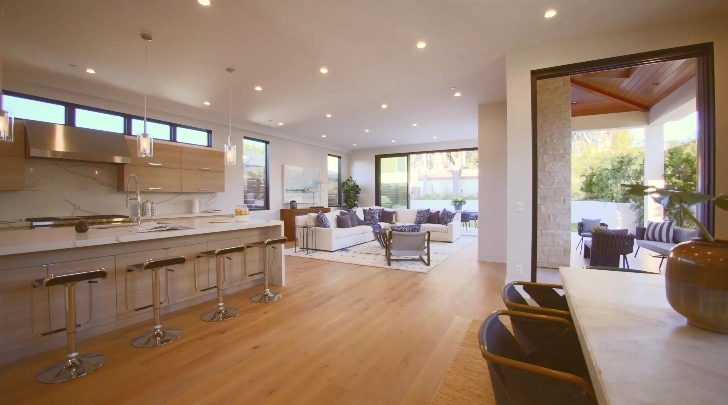 Open-concept modern living area with a kitchen island and bar stools, white sectional sofa with patterned pillows, and a patio with outdoor seating.