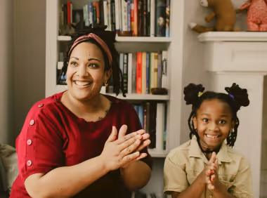 Smiling woman and young girl sitting indoors, clapping hands together with a bookshelf and fireplace behind them.
