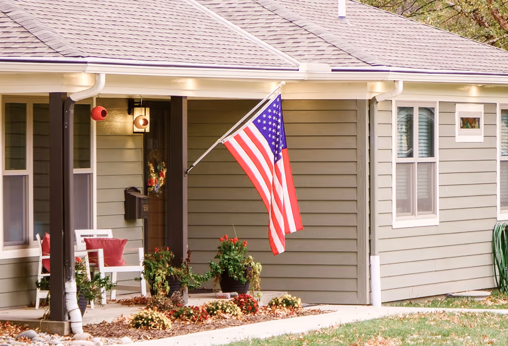 Front porch of a gray house with an American flag, white chairs with red pillows, and flower beds.