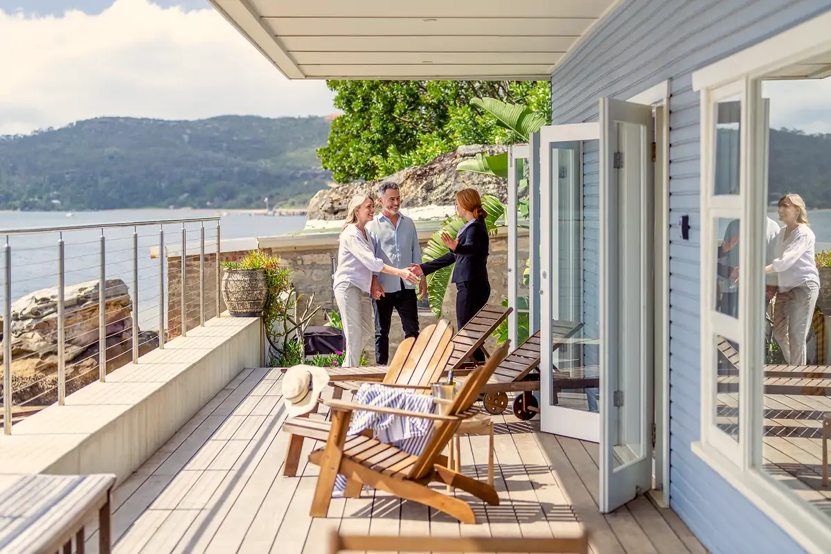 Couple shaking hands with a real estate agent on a wooden deck overlooking a scenic waterfront and hills.