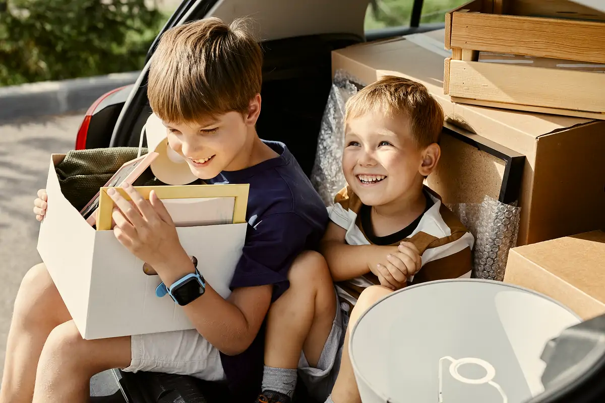 Two young boys sitting in the back of a car surrounded by moving boxes, one holding a box of household items.