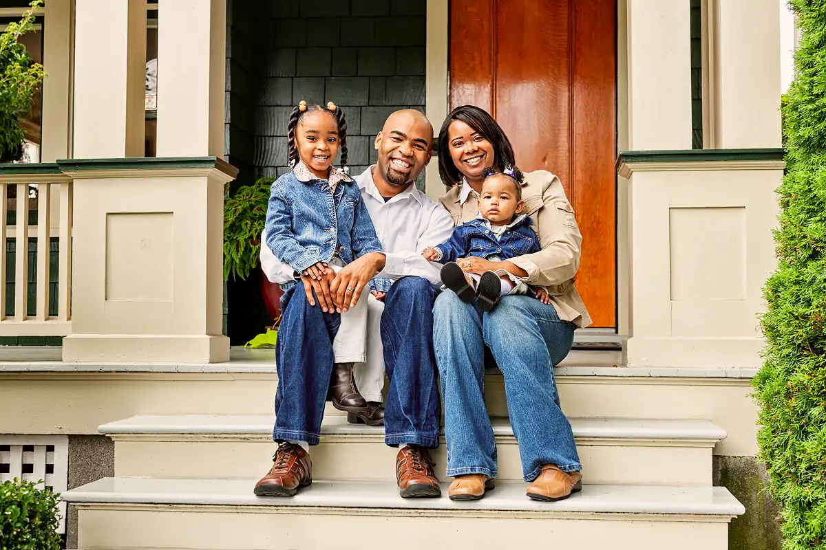 Smiling African American family of four sitting on the front steps of a house with a wooden door.