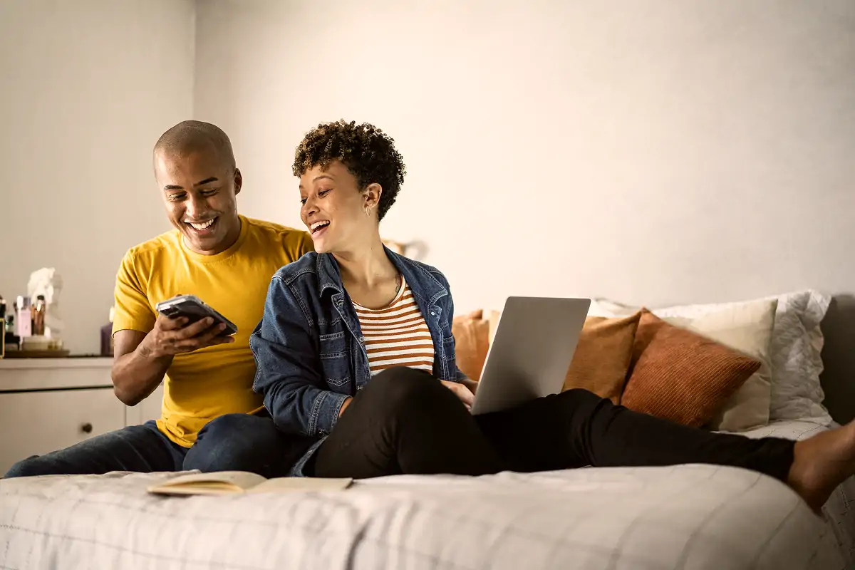 Smiling couple relaxing on bed with laptop and smartphone, looking at phone screen together.