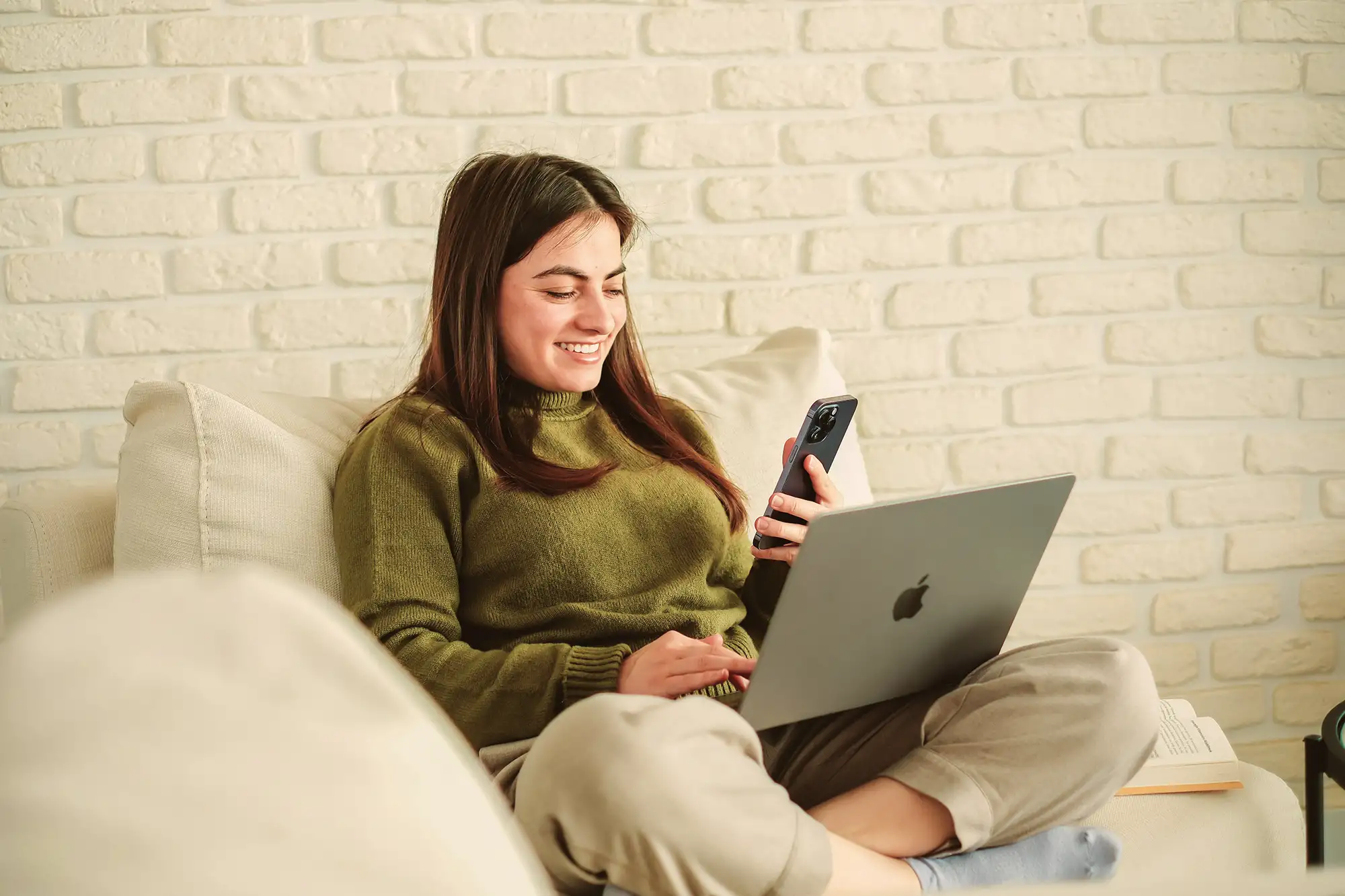 Smiling woman sitting cross-legged on a couch using a laptop and holding a smartphone.