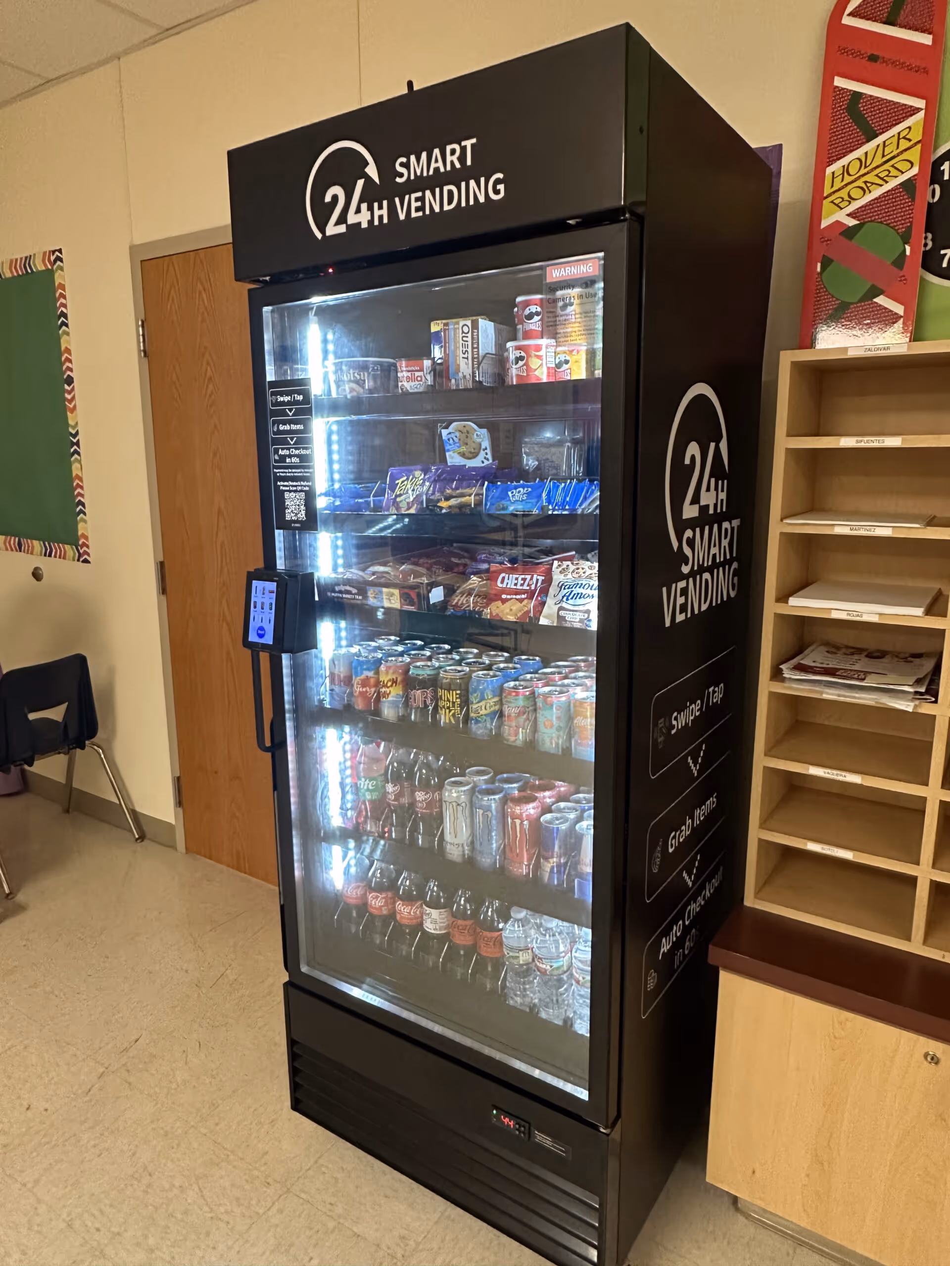 Black smart vending machine labeled 24H Smart Vending filled with various snacks and beverages including chips, cookies, soda cans, and bottled water.
