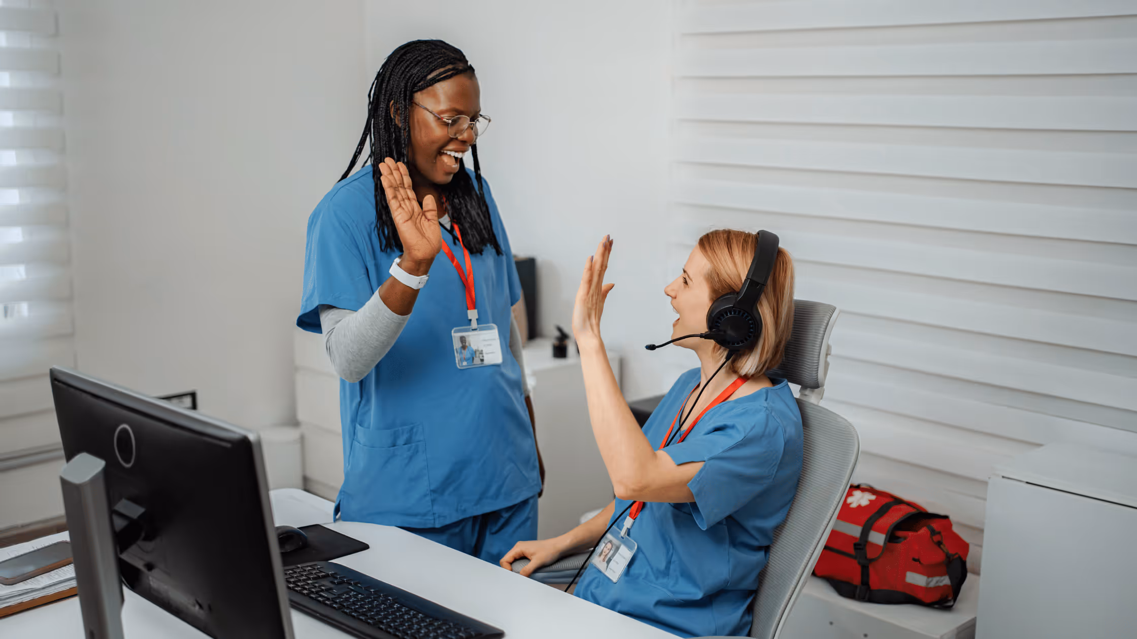 Two healthcare workers in blue scrubs giving each other a high-five in an office setting.