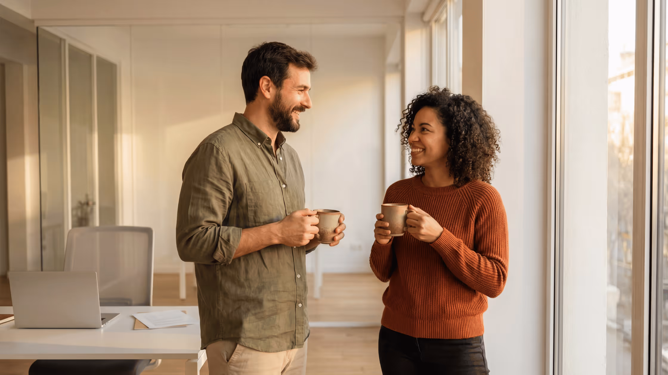 Two colleagues smiling and talking while holding coffee mugs in a bright office near large windows.