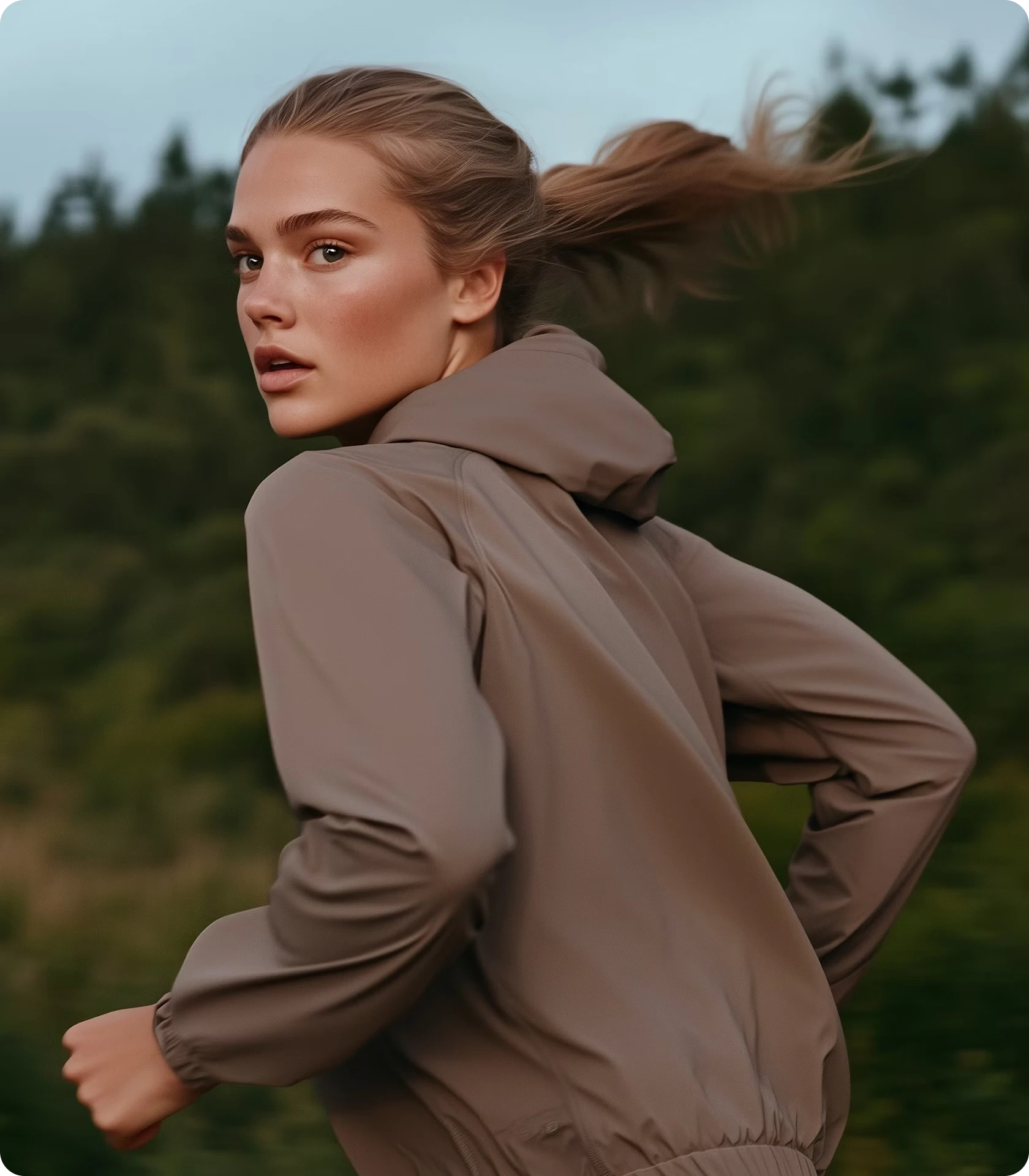 Young woman with ponytail wearing a brown jacket, running outdoors with a blurred green forest background.