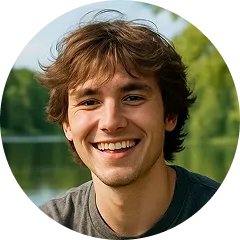 Smiling young man with tousled brown hair outdoors near a body of water and greenery.
